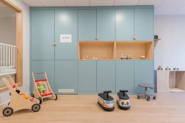 Salle d'activités de la crèche Les Bébés Explorateurs Cormeilles-en-Parisis (95240) – grande armoire murale bleu canard avec niches bois, porteurs panda et bus, poussette poupée bois et étagère à jouets