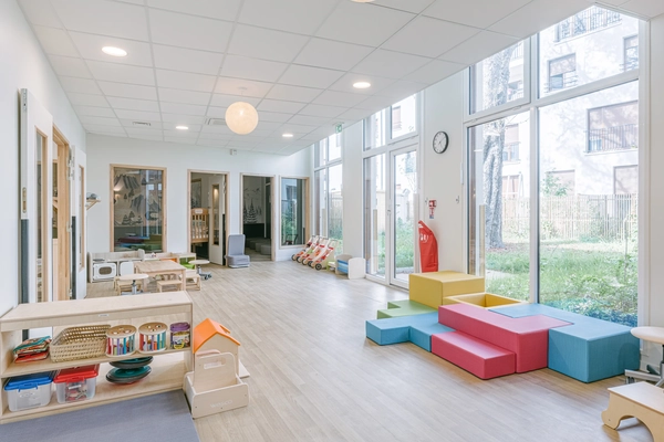 Salle de jeux de la crèche Les Bébés Explorateurs Montreuil (93100) – modules mousse colorés bleu, jaune et rouge, étagères Montessori, grandes baies vitrées ouvrant sur le jardin
