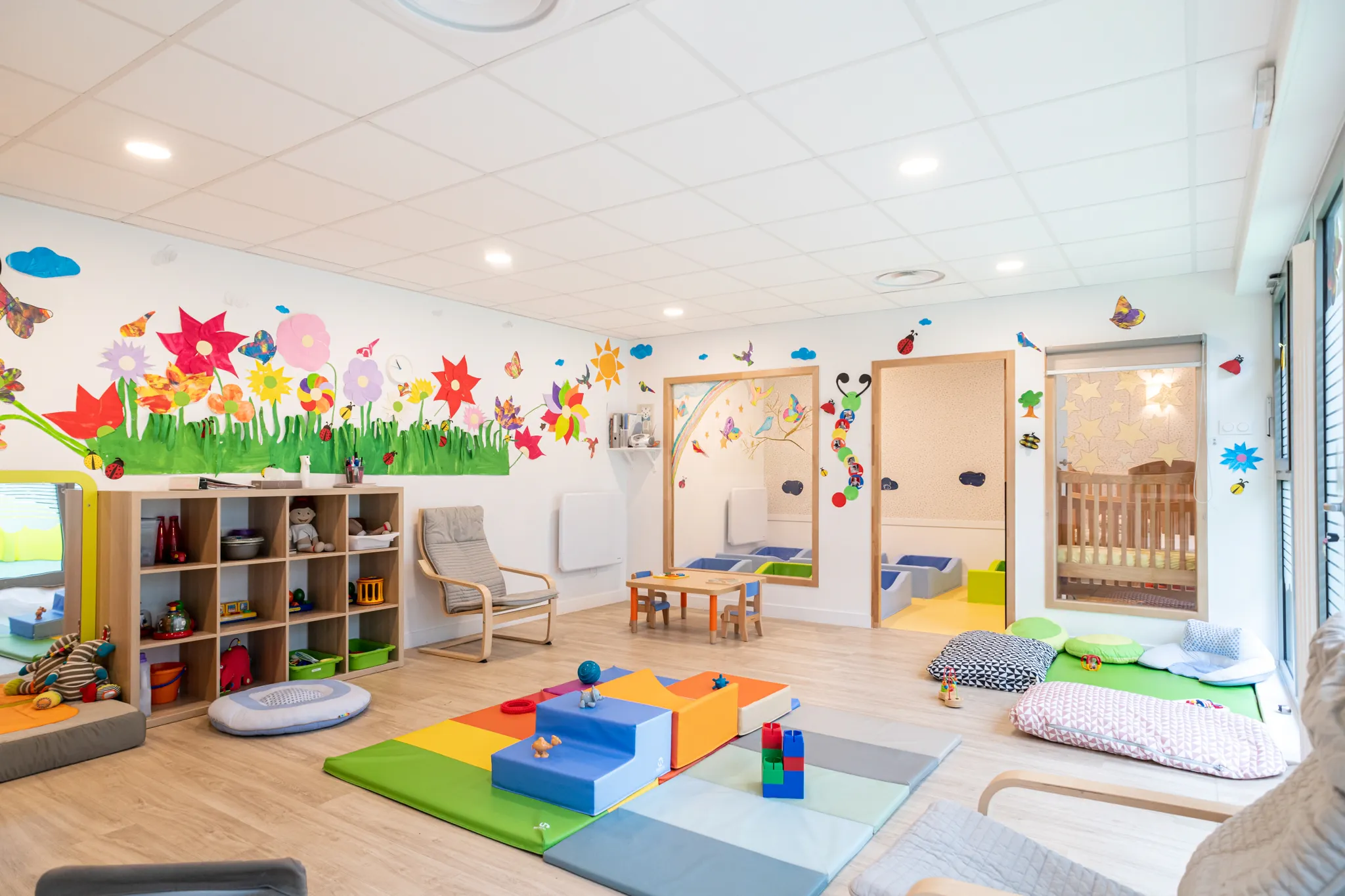 Salle d'éveil de la crèche Les Bébés Explorateurs Nogent Baltard (94130) – grande fresque florale colorée au mur, modules de motricité multicolores, fenêtre vitrée sur le dortoir et coin lecture